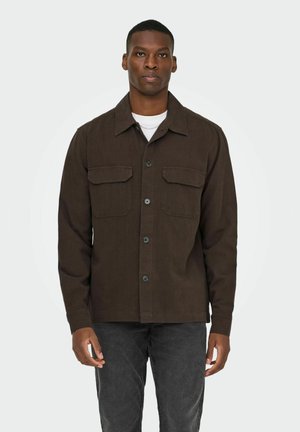 Mann står iført en mørk brun knappesjakke over en hvit skjorte og mørkegrå bukser mot en ensfarget lys bakgrunn.