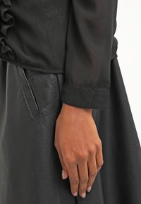 Blouse en satin noir avec détails de manches à volants, associée à une jupe en cuir noire. Caractérisée par une texture douce et un design élégant et moulant.