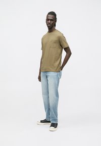 Olívazöld rövid ujjú póló zsebbel, világoskék farmernadrággal és fekete, fehér talpú tornacipővel párosítva.