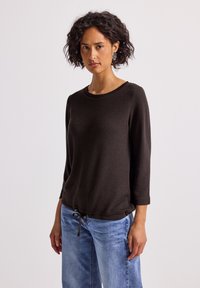 Brązowy sweter z dzianiny z rękawami o długości trzech czwartych, okrągłym dekoltem i ściągaczem na dole. Połączony z jasnoniebieskimi spodniami z szerokimi nogawkami.