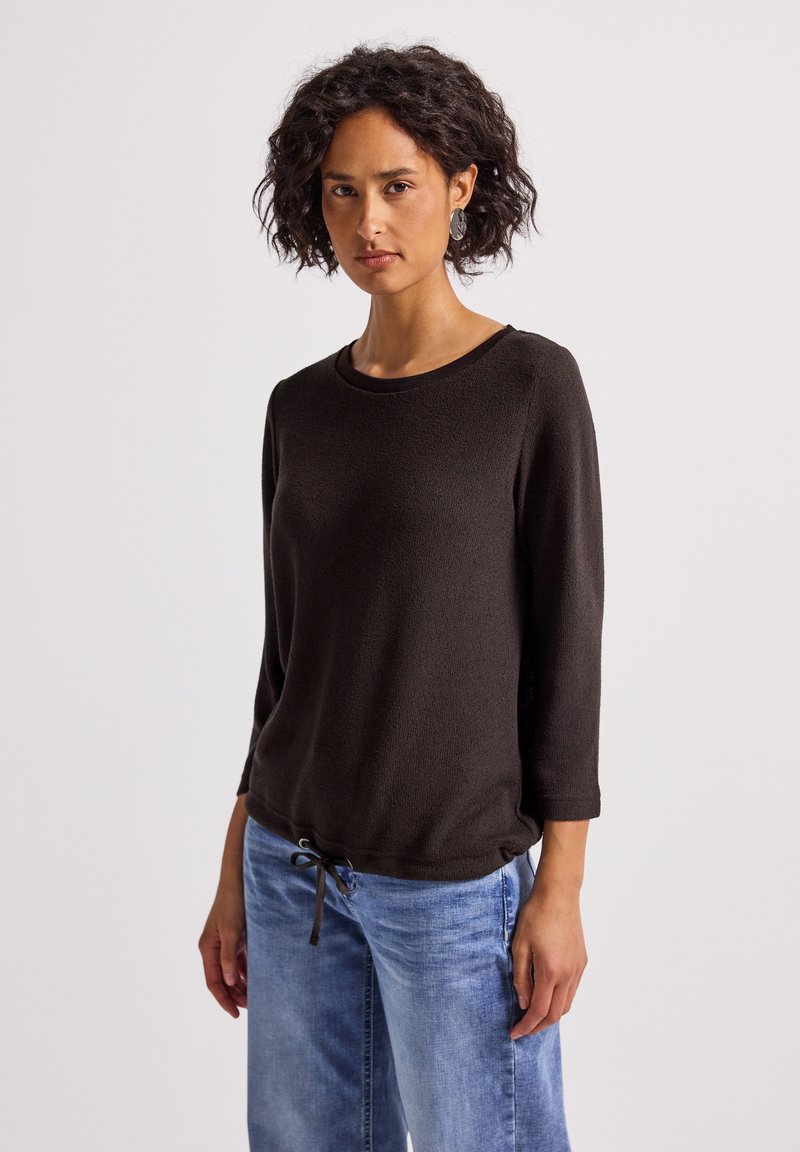 Brązowy sweter z dzianiny z rękawami o długości trzech czwartych, okrągłym dekoltem i ściągaczem na dole. Połączony z jasnoniebieskimi spodniami z szerokimi nogawkami.