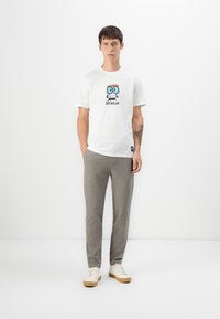 Joven con cabello corto que está de pie vistiendo una camiseta blanca de dibujo animado con la palabra "Genius", pantalones grises, zapatillas blancas y un tatuaje visible en el antebrazo.