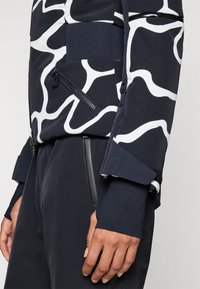 Marineblauer Blazer mit weißem abstrakten Muster, ausgestattet mit einem gerippten Bund, Reißverschlusstaschen und strukturierten Bündchen, gefertigt aus einem glatten synthetischen Material.