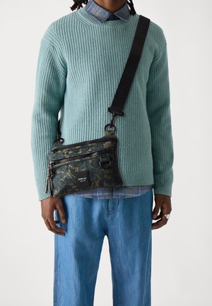 Czarna i zielona torba na ramię z podwójnymi zamkami błyskawicznymi, regulowanym paskiem i naszywką z logo. Noszona na jasnoniebieskim swetrze w prążki.