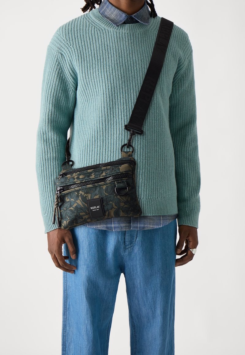 Schwarz-grün gemusterte Umhängetasche mit zwei Reißverschlüssen, verstellbarem Riemen und Markenpatch. Getragen über einem hellblauen, gerippten Pullover.