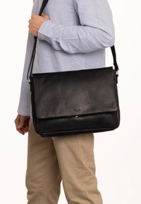 Black leather messenger bag with a smooth texture, magnetic flap closure, adjustable strap, and a subtle logo embossed on the front.