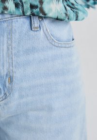 Vaaleansiniset farkut, joissa on klassinen leikkaus, etutaskujen saumat ja näkyvät tikkauslinjat. Kuvioitu vihreä ja musta paita peittää osittain vyötärön.