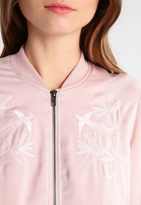 Lichtroze bomberjack met geborduurde vogels en loof, rits aan de voorkant en een zachte textuur. Ribbelkraag en manchetten.