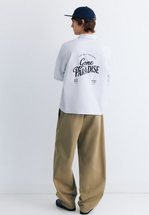 Jeune personne portant une casquette marine, une chemise légère et ample avec le texte "Gone Paradise" au dos, et un pantalon large beige sur un fond blanc uni.