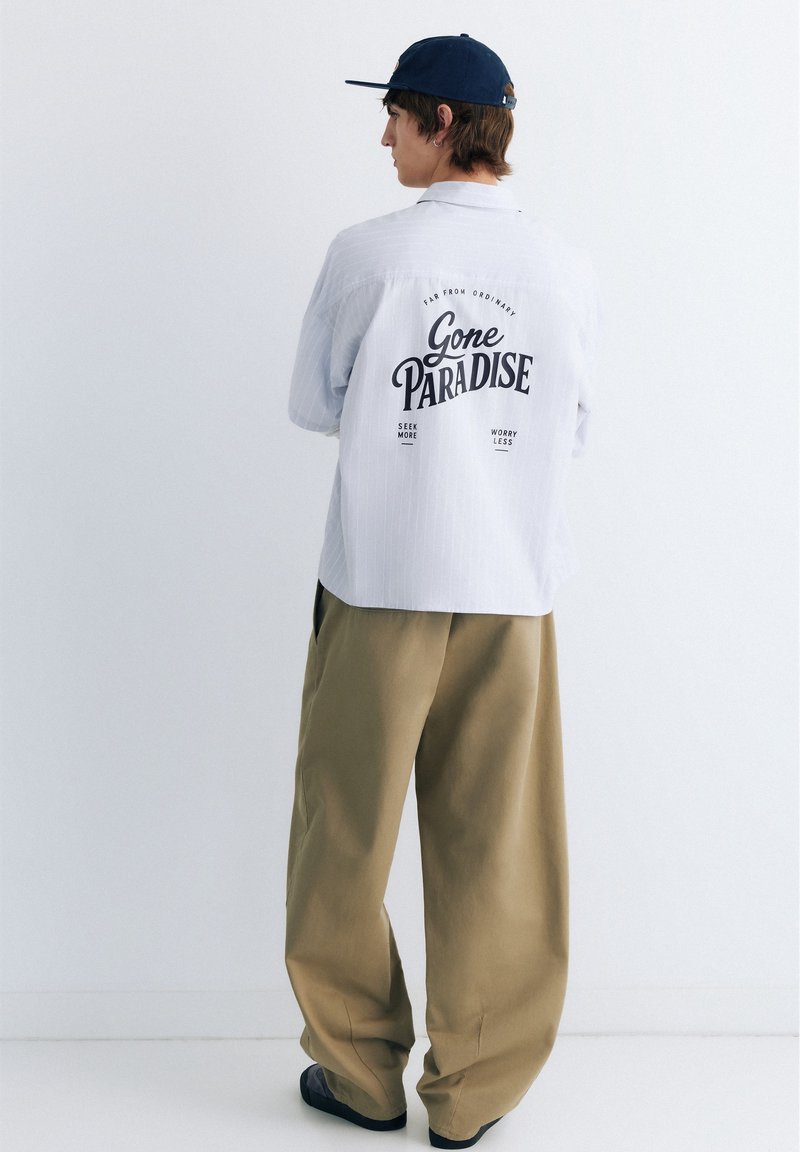 Jeune personne portant une casquette marine, une chemise légère et ample avec le texte "Gone Paradise" au dos, et un pantalon large beige sur un fond blanc uni.