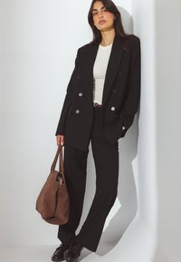 Kobieta w czarnym garniturze w prążki i białej bluzce, trzymająca brązową zamszową torebkę, stojąca oparta o białą ścianę w swobodnej pozie.