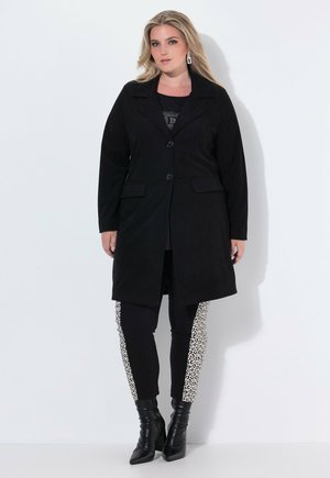 Cappotto in corduroy nero con colletto a mostrine e un solo bottone. Indossato su una maglietta nera e leggings neri con pannelli laterali in stampa leopardata. Stivali neri con tacco.