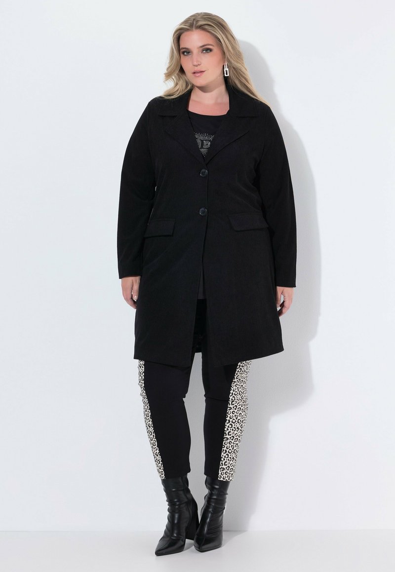 Cappotto in corduroy nero con colletto a mostrine e un solo bottone. Indossato su una maglietta nera e leggings neri con pannelli laterali in stampa leopardata. Stivali neri con tacco.