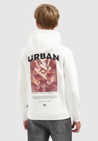 Blond persoon in een witte hoodie met de capuchon op, voorzien van een geometrisch stedelijk ontwerp en tekst op de achterkant, gecombineerd met donkergrijze jeans.