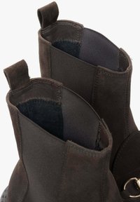 Brązowe botki zamszowe z elastycznymi bokami, miękką podszewką i pętelkami do zakładania. Charakterystyczny metalowy pierścień na zewnętrznej stronie. Gumowa podeszwa.