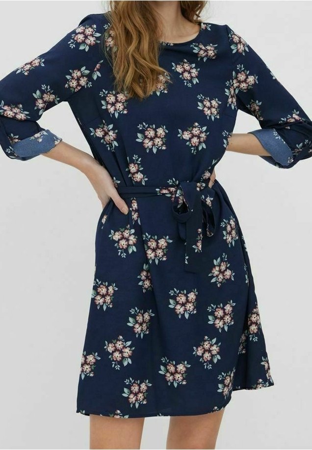 Robe bleu marine mi-longue avec motif floral rose, manches longues retroussées et ceinture nouée à la taille.