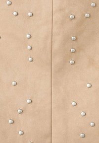 Tela de ante beige con tachuelas metálicas plateadas espaciadas uniformemente en un patrón decorativo. La textura es suave con un ligero brillo.