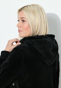 Sort hoodie lavet af blødt stof, med en hætte og tekstureret overflade. Modellen vises fra siden, med hånden på kraven.