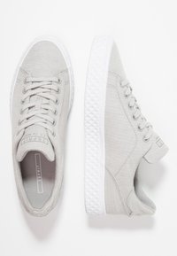 Jasnoszare płócienne trampki z teksturowaną powierzchnią, sznurowanym fasonem i białą gumową podeszwą z diamentowym wzorem wzdłuż krawędzi.