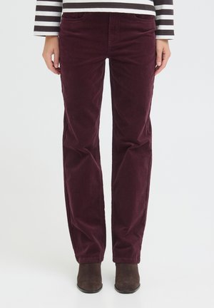 Personne portant un pantalon en velours côtelé violet foncé et des bottines en daim marron, visible de la taille aux pieds sur un fond uni.