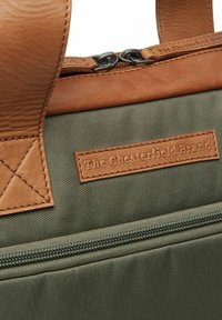 Sac khaki avec des accents en cuir, doté d'une fermeture éclair et d'une étiquette cousue. Cuir texturé et tissu durable dans des tons de vert et de marron.