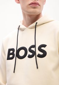 Jaunas vyras, apsirengęs kreminės spalvos džemperiu su juodomis raištelėmis ir dideliu juodu "BOSS" užrašu ant krūtinės, priešais paprastą baltą foną.