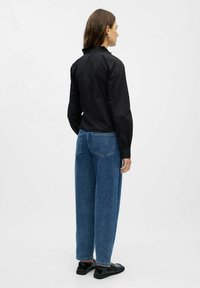 Frau in einer schwarzen Bluse mit langen Ärmeln, blauen hochtaillierten, geraden Jeans und schwarzen Slippern, die mit dem Rücken zu einem weißen Hintergrund steht.