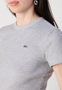 Jauna moteris su tamsiais plaukais dėvi prigludusią pilką raukšlėtą marškinėlių, ant kurios yra mažas žalias krokodilo logotipas, ir didelius sidabrinius auskarus.