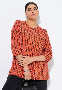 Nainen, joka wearing punasävyistä 3/4-hihasta puseroa, mustia housuja, hopeisia korviksia ja pitää mustaa laukkuua valkoista taustaa vasten.