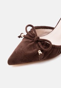 Καφέ γυναικείο παπούτσι από σουέτ με μυτερή μύτη, μάζεμα στις άκρες και δεμένη φιόγκο μπροστά με μικρές μεταλλικές χάντρες.
