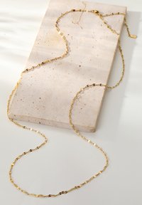 Goldkettchen mit verlängerten, verlinkten Elementen auf einer strukturierten, hellen Steinoberfläche, mit einemverschluss und einem kleinen Tag-Detail.
