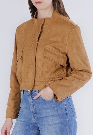 Kvinde iført en kort, beige ruskindjakke med klaplommer og blå højtaljede jeans, med hånden i lommen, foran en ensfarvet baggrund.