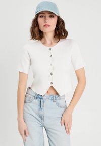 Crop top blanc à manches courtes et col rond, doté de boutons en forme de cœur sur le devant. Associé à un jean en denim bleu clair.