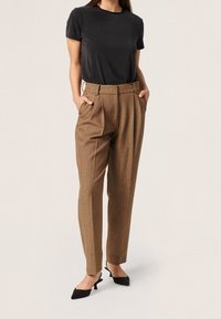 Brązowe spodni w kratę o dopasowanym kroju z wysokim stanem, plisowanym przodem i bocznymi kieszeniami, w połączeniu z dopasowaną czarną koszulką i spiczastymi czarnymi butami.