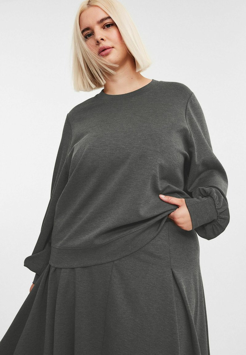 Kobieta z blond bobem, ubrana w luźną, ciemnoszaro-skalną bluzkę i pasującą spódnicę, trzymająca materiał bluzki jedną ręką.