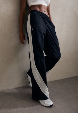 Persona con pantalones deportivos en blanco y negro, top deportivo blanco y zapatillas deportivas blancas, de pie contra una pared lisa.