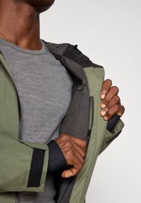 Zöld dzseki szürke bélésse, cipzáras zsebbel és tépőzáras díszítéssel az ujjakon, egy szürke póló felett. Texturált szövet.