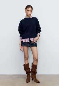 Pull en maille bleu marine superposé sur une chemise rayée rouge et blanche, associé à une mini-jupe en jean bleu et des bottines à talons molles en daim marron.