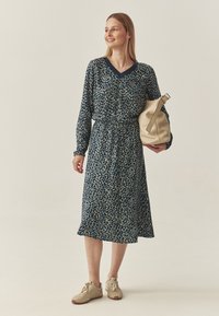 Rochie bleumarin cu un model floral, de tip v-neck, cu mâneci lungi. Rochia are o talie elastică și o fustă fluidă. Modelul ține o geantă bej.