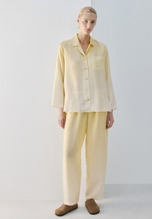 Woman standing against a plain wall wearing loose pale yellow button-up shirt, matching wide-leg pants, and brown slip-on shoes.