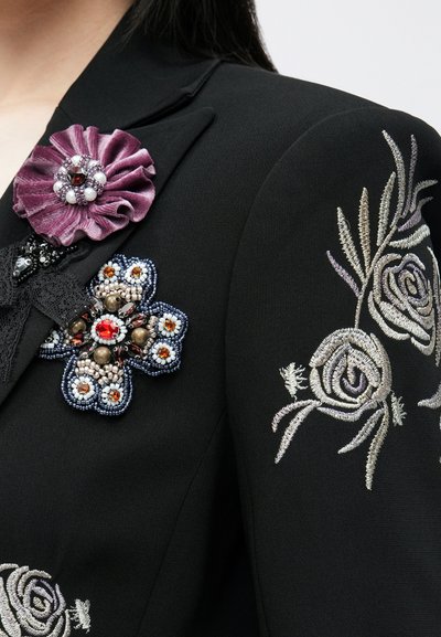 Fekete dzseki aprólékos virágos hímzéssel. Díszes brossok lila, kék és ezüst árnyalatú szövetből és gyöngyökből.