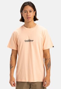 Camiseta corta de manga en color melocotón claro, hecha de algodón. Presenta un estampado de logo negro "Quiksilver" en el pecho. Diseño de cuello redondo.