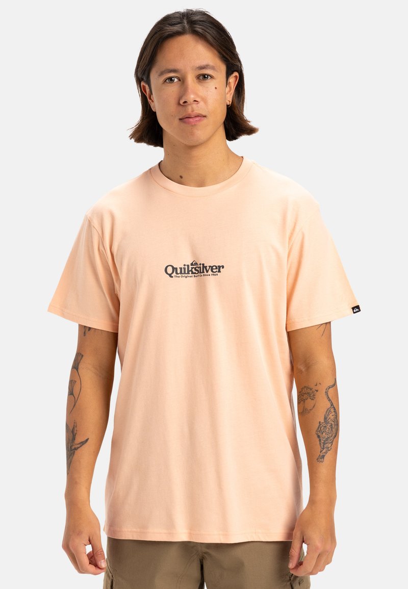 Camiseta corta de manga en color melocotón claro, hecha de algodón. Presenta un estampado de logo negro "Quiksilver" en el pecho. Diseño de cuello redondo.