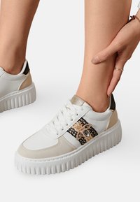 Sneaker blanc avec des accents beiges et une bande texturée noire et dorée. Présente une semelle épaisse et crantée ainsi qu'un matériel décoratif sur le côté.