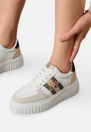 Sneaker blanc avec des accents beiges et une bande texturée noire et dorée. Présente une semelle épaisse et crantée ainsi qu'un matériel décoratif sur le côté.