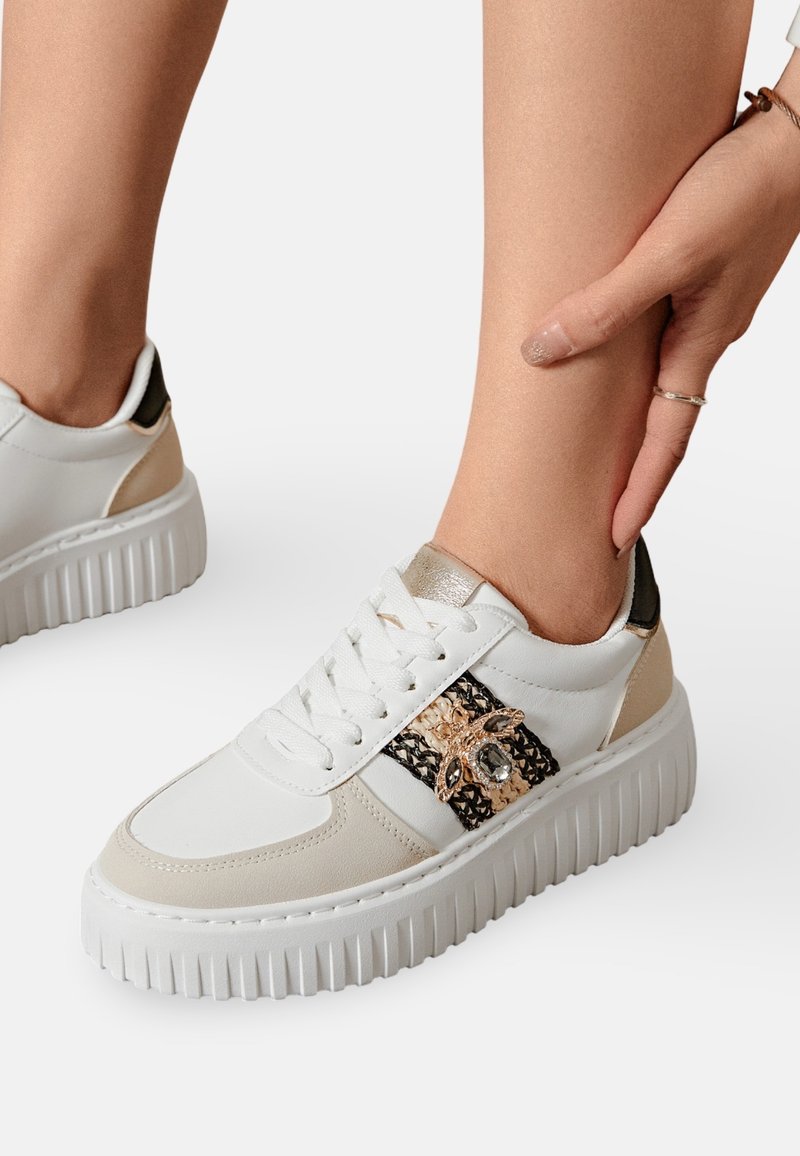 Sneaker blanc avec des accents beiges et une bande texturée noire et dorée. Présente une semelle épaisse et crantée ainsi qu'un matériel décoratif sur le côté.