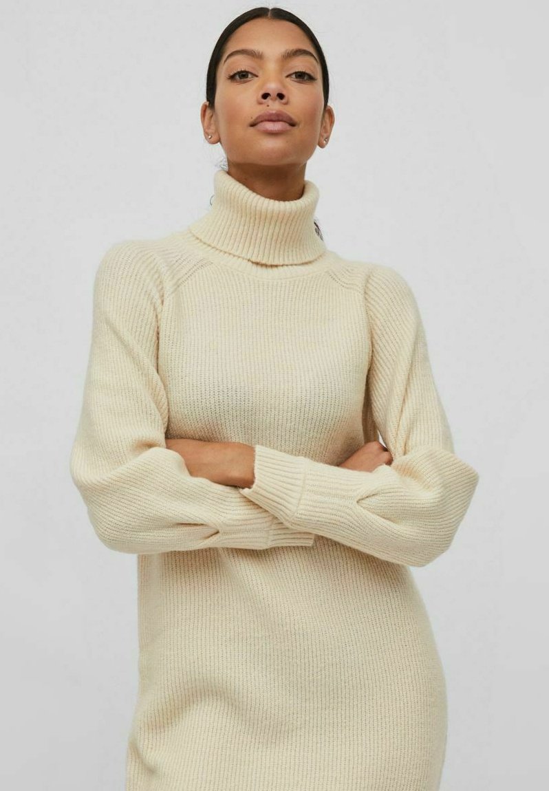 Robe pull tricotée de couleur crème avec un col roulé haut, texture côtelée, manches longues et coupe légèrement oversized.