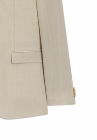 Manga y detalle lateral de chaqueta de traje beige claro con bolsillo con solapa y tres pequeños botones en el puño, exhibida sobre fondo blanco.