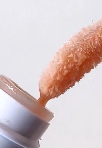 Applicateur de gloss à lèvres couvert de gloss de couleur pêche, soulevé au-dessus d'un contenant de gloss ouvert sur fond blanc.