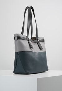 Borsa a spalla in pelle bicolore grigia e blu scuro con doppi manici neri e dettagli in fibbia, esposta su un piedistallo bianco.